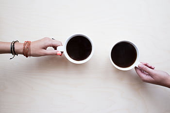 Two female hands holding coffee mugs on a light wooden table, symbolizing friendship and relaxation. HD wallpaper