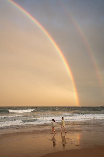 Two children walking along a beach under a vibrant rainbow during twilight. HD phone wallpaper