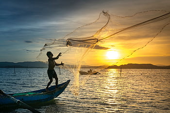A fisherman skillfully casts a net at sunrise, silhouetted against a golden sky over a calm lake. HD wallpaper