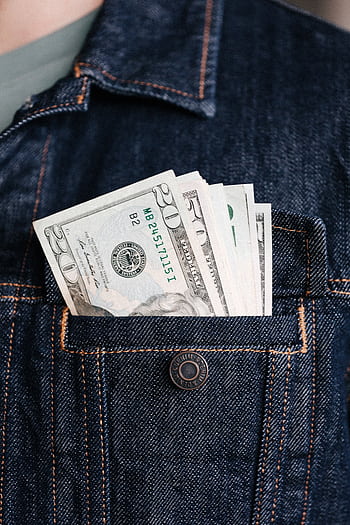 Close-up of US dollar bills in a denim jacket pocket, symbolizing wealth and finance. HD phone wallpaper