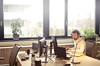 A businessman sits at a desk using multiple computers and a headset in a well-lit modern office. HD wallpaper