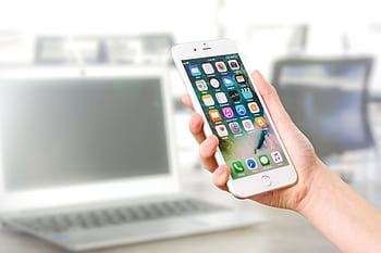 Close-up view of a smart displaying apps, held by a hand, with a blurred laptop in the background. HD wallpaper