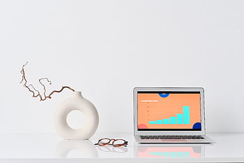 A minimalist workspace with a laptop, ceramic vase, and eyeglasses on a white desk. HD wallpaper