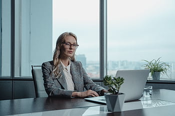 A confident businesswoman in a grey suit working on a laptop in a modern office setting. HD wallpaper