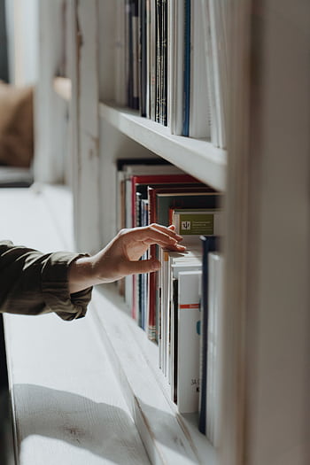 A hand reaches out to select a book from a well-arranged indoor bookshelf. HD phone wallpaper