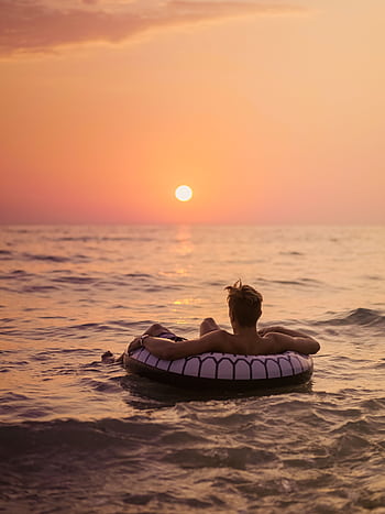 Man enjoying a serene ocean sunset while floating on an inflatable ring, capturing tranquility and leisure. HD phone wallpaper