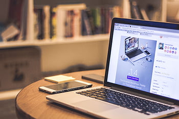 A cozy modern workspace featuring a laptop displaying a website, a smart, and books on shelves. HD wallpaper