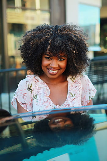 Smiling woman with natural afro hairstyle outdoors in a floral dress, capturing joy on a sunny day. HD phone wallpaper