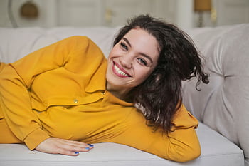 A joyful woman in a yellow outfit smiling while lying on a sofa at home. HD wallpaper