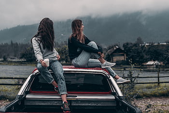 Two young women sitting on a car, enjoying the serene mountain landscape in Romania. HD wallpaper