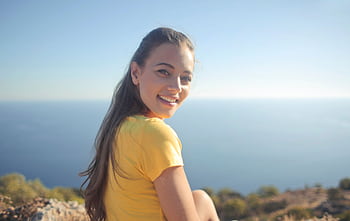 Young woman in a yellow shirt smiles by the sea, enjoying the outdoor view. HD wallpaper