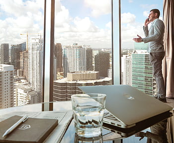 Businessman talking on smart by window with city skyline view, laptop and glass in foreground. HD wallpaper