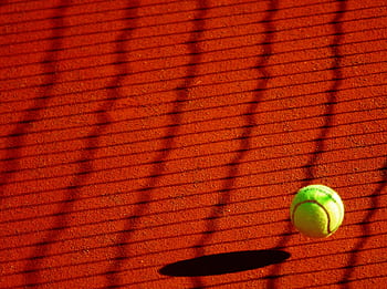 A vibrant tennis ball casting a shadow on a sunlit clay court with net shadows. HD wallpaper