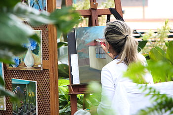 A woman enjoys painting in a lush outdoor garden, surrounded by vibrant artwork. HD wallpaper