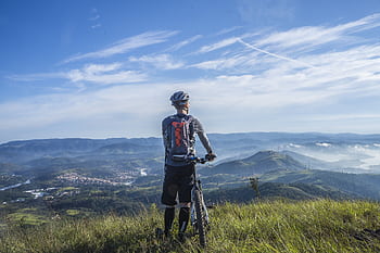 A cyclist stands on a hill, gazing at a stunning mountain landscape on a bright sunny day. HD wallpaper