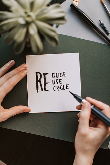Close-up of hands writing a motivational 'Reduce Reuse Recycle' note on paper with a pen, surrounded by office items. HD phone wallpaper
