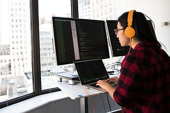 Woman programming on a laptop at a standing desk in an office with large windows. HD wallpaper