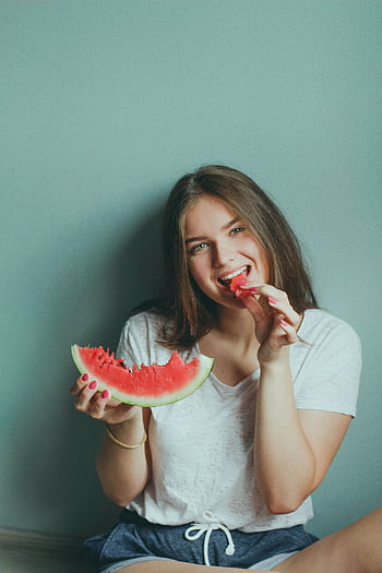 Smiling young woman sitting indoors enjoying a fresh slice of watermelon. HD phone wallpaper