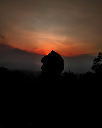 A serene silhouette of a person wearing a hijab against a vibrant sunset sky in a rural area. HD phone wallpaper
