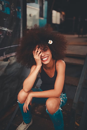 Cheerful woman wearing ripped jeans and curly hair with flower, enjoying a sunny day outdoors. HD phone wallpaper