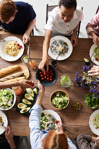 A group of people sharing a vibrant meal with salads, pasta, and fresh vegetables, promoting togetherness. HD phone wallpaper
