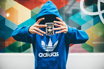 A mysterious magician in a blue hoodie performing a card trick with a colorful geometric background. HD wallpaper