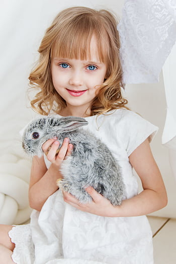 Charming young girl smiling while holding a fluffy grey bunny, radiating joy and innocence indoors. HD phone wallpaper