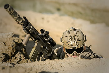 A soldier with tactical gear hiding in the desert, poised for action. HD wallpaper