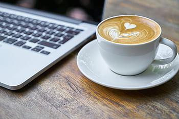 A cup of latte with intricate art beside a laptop on a wooden table, depicting a cozy work setting. HD wallpaper