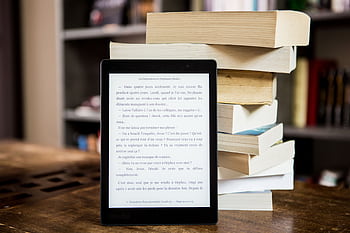 E-reader with text displayed next to a stack of physical books on a wooden table. HD wallpaper