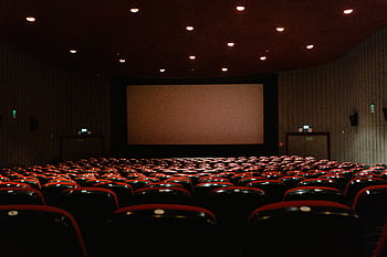 A dimly lit empty cinema theater with rows of red chairs facing a projector screen. HD wallpaper
