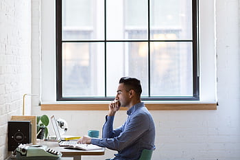 Focused male professional working on a laptop at an office desk by a large window. HD wallpaper