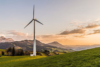 Wind turbine amid rolling hills and mountains at sunset, symbolizing renewable energy and sustainability. HD wallpaper