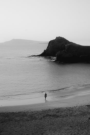 A lone person walks along a tranquil beach with rocky cliffs, captured in monochrome. HD phone wallpaper