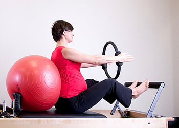 A pregnant woman in a red top practicing Pilates using a fitness ring and exercise ball in a gym setting. HD wallpaper