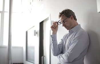 Thoughtful man in a bright room holding his glasses while leaning against a wall. HD wallpaper