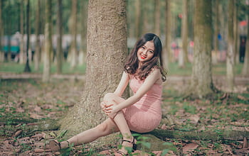 Smiling young woman sits against a tree in a sunny park setting, enjoying leisure time. HD wallpaper
