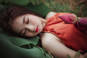 A serene woman in an orange dress lying on green leaves with lotus flowers. HD wallpaper