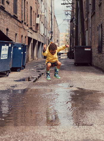 A young boy in a yellow outfit jumps over a puddle in an urban alley. Vibrant and playful moment. HD phone wallpaper