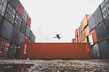 A person leaps dramatically above stacked shipping containers, showcasing dom and industrial energy. HD wallpaper