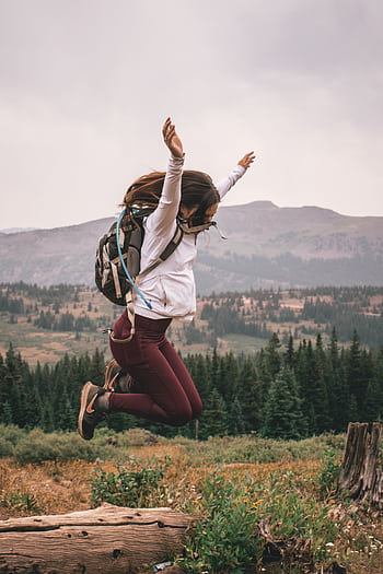 Joyful young woman leaps over a log in a scenic mountainous forest setting, embodying outdoor adventure. HD phone wallpaper