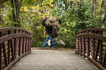 A blonde girl jumps exuberantly on a wooden bridge surrounded by autumn foliage, capturing the essence of joy and dom. HD wallpaper