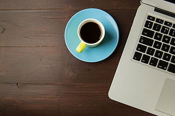 A top view of a coffee cup on a saucer next to a laptop on a wooden desk, ideal for work-from-home themes. HD wallpaper