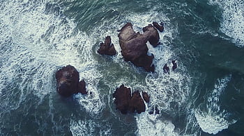 Dynamic aerial view of ocean waves crashing against rocky structures, captured from above. HD wallpaper