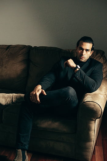 Elegant businessman in a suit relaxing on a brown couch with a serious expression. HD phone wallpaper