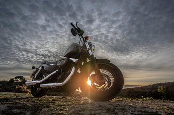 A striking of a motorcycle parked on a dirt road with a dramatic sky at sunset, capturing the sun glare. HD wallpaper