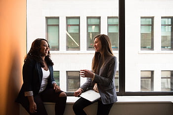 Two businesswomen enjoying a conversation by a large office window. HD wallpaper