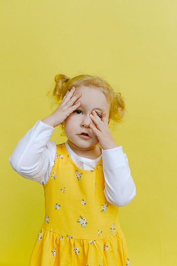 Adorable toddler with hands on face wearing a yellow floral dress against a yellow background. HD phone wallpaper
