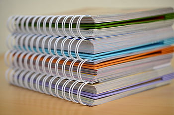 A colorful stack of spiral-bound notebooks neatly placed on a wooden desk. Ideal for educational themes. HD wallpaper