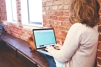 A woman is using a laptop for remote work while sitting indoors against a brick wall. HD wallpaper
